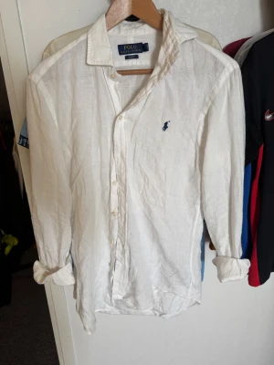 Vit linneskjorta från Polo Ralph Lauren - Klassisk vit skjorta i linne från Polo Ralph Lauren med blå broderad logga på bröstet. Skjortan har lång ärm, knappar framtill och en avslappnad passform. Perfekt för en stilren och enkel look.