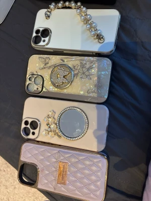 Fyra lyxiga mobilskal med pärlor & glitter iphone 15 - Säljer fyra snygga mobilskal till iPhone15, alla med unika detaljer som pärlor, blommor, spegeleffekt och quiltad design. Färgerna går i vitt, beige och pärlemor med inslag av guld och silver. Skal med ringhållare, pärlhandtag och blommor – perfekta för dig som gillar glam och elegans.