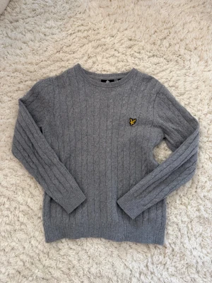 Grå kabelstickad tröja Lyle & Scott - Snygg grå kabelstickad tröja från Lyle & Scott med rund halsringning och klassisk gul logga på bröstet. Tröjan har långa ärmar och ribbade muddar vid ärmslut och nederkant. Perfekt för lager på lager och enkel att matcha. Storlek S men sitter lite mindre!