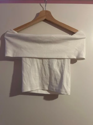 Vit offshoulder topp Gina Tricot - Säljer en vit offshoulder topp från Gina Tricot i mjuk bomull. Toppen har en bred kant som vilar över axlarna och är axelbandslös för en clean och stilren look. Perfekt att styla med höga jeans eller kjol.