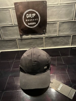 Svart keps från BOSS - helt ny snygg svart keps från BOSS med klassisk böjd skärm och justerbar rem bak. Kepsen har en diskret metallogo framtill och är tillverkad i ett slitstarkt tyg som passar perfekt till streetwear eller casual outfits.