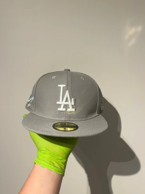 Grå LA Dodgers keps från New Era - Snygg grå keps från New Era med LA Dodgers-logga framtill i vitt och broderad 1988 World Series-detalj på sidan. Klassisk 59FIFTY-modell med platt skärm och strukturerad passform. Perfekt för dig som gillar sportig streetstyle.