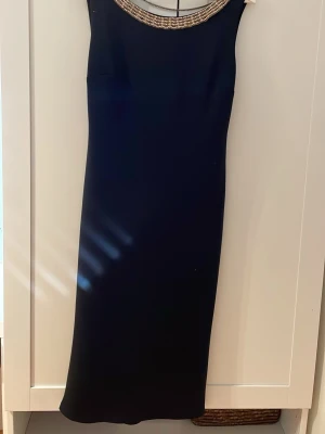 Svart långklänning med gulddetaljer - Elegant svart långklänning med guldfärgade pärldetaljer runt halsringningen och dekorativa kedjor i ryggen. Klänningen har ärmlös design och rak passform, perfekt för en stilren look. Materialet känns mjukt och följsamt.