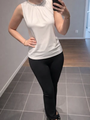 Vit ärmlös topp med pärldetaljer - Snygg vit ärmlös topp med rund halsringning dekorerad med pärlor. Toppen har en rak passform och är tillverkad i ett mjukt material som känns skönt mot huden. Perfekt att styla med både jeans och kjol för en fräsch look.