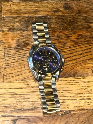 Michael Kors klocka i guld och silver - Snygg Michael Kors klocka med länkarmband i både guld och silver. Urtavlan är mörk blå med romerska siffror och flera små urtavlor för extra funktioner. Klockan har en klassisk och lyxig design som passar perfekt till din stil. Boettstorlek: 45 mm Bandbredd: 20 mm Passar handled ca 16–20 cm  Storlek kan ändras om man har länkar