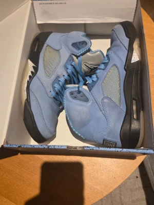 Air Jordan 5 Retro UNC blå sneakers - Snygga Air Jordan 5 Retro UNC sneakers i ljusblå mocka med svarta och vita detaljer. Skorna har meshpaneler på sidorna, klassisk hög siluett och matchande blå skosnören. Perfekta för dig som vill sticka ut med stilrena och ikoniska sneakers.
