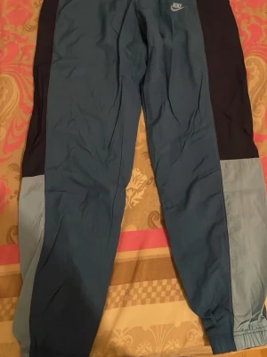 Nike blå och svarta mjukisbyxor - Säljer ett par snygga mjukisbyxor från Nike i olika blå nyanser och svart. Byxorna har elastiska muddar vid bensluten och en liten Nike-logga på höften. Tillverkade i ett lätt och bekvämt material, perfekta för chill eller träning.