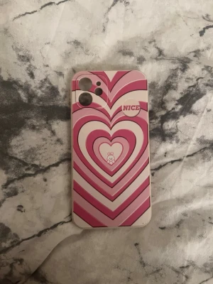 Rosa hjärtmönstrat mobilskal för iPhone 12 - Säljer ett mobilskal med rosa och vita hjärtan i retrostil. Skalet har texten 'NICE' och en liten söt björn i mitten. Tillverkat i plast och passar iPhone 12 . Perfekt för dig som vill ha ett gulligt och färgglatt skal. Några få repor som knappt är synliga.