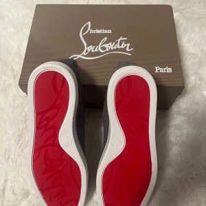 Gråa sneakers från Christian Louboutin med röd sula - Säljer ett par gråa sneakers från Christian Louboutin med ikonisk röd sula. Skorna har snörning, rund tå och är tillverkade i mocka med vita detaljer på sulan. Perfekta för dig som vill sticka ut med exklusiva och stilrena skor.
