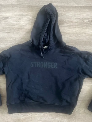 Svart hoodie från Stronger - Svart cropped hoodie från Stronger med huva och dragsko. Tryckt logga på bröstet i samma färg för en clean look. Perfekt till jeans eller tights. Mjuk bomullskänsla och avslappnad passform.