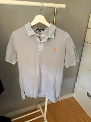 Ljusgrå pikétröja från Ralph Lauren - Säljer en klassisk ljusgrå pikétröja från Ralph Lauren med korta ärmar och den ikoniska broderade loggan i orange på bröstet. Tröjan har krage och knäppning framtill, perfekt till jeans eller shorts.