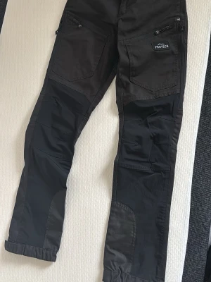 Svarta jobbbyxor från Protech - Svarta jobbbyxor från Protech med flera praktiska fickor och dragkedjor. Byxorna har förstärkta partier på knän och benslut samt justerbara benslut med kardborre. Perfekta för en aktiv livsstil och outdoor-äventyr.