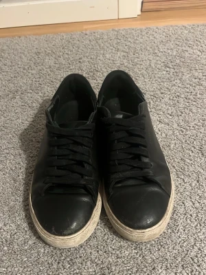 Svarta sneakers från Axel Arigato - Stilrena svarta sneakers i läder från Axel Arigato med svart snörning och vit sula. Klassisk låg modell som passar till det mesta och har diskret logga på sulan. Perfekta för dig som gillar minimalistisk och clean stil.