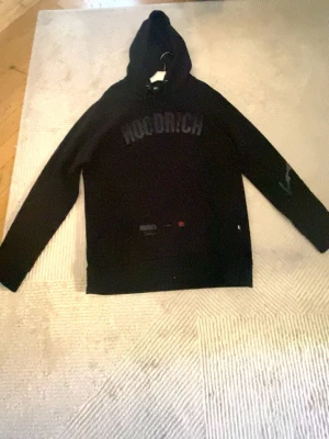  Hoodrich hoodie  - Svart hoodie från Hoodrich med stor logga på bröstet och tryck på ryggen. Klassisk modell med huva och långa ärmar. Snygga detaljer på ärmen och framtill. Perfekt för dig som gillar streetwear och vill ha en stilren look. Passar runt 175-185 som storlek M.
