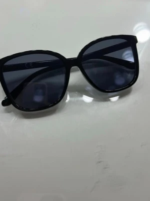 Svarta solglasögon med bred båge - Stilrena svarta solglasögon med bred båge och mörka glas. Klassisk och tidlös design som passar till de flesta outfits. Perfekta för soliga dagar när du vill ha en cool look. Vet tyvärr inte vart de är ifrån. 