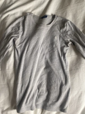 Ljusgrå långärmad topp Polo Ralph Lauren - Ljusgrå långärmad topp från Polo Ralph Lauren med rund halsringning och diskret broderad logga på bröstet. Tillverkad i mjukt bomullsmaterial som känns skönt mot huden. Enkel och stilren design som passar till det mesta.