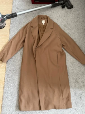 Kamelbrun klassisk kappa från H&M - Stilren kamelbrun rock från H&M med dubbelknäppning och klassisk krage. Lång modell med raka ärmar och slät yta, perfekt för en trendig och tidlös look. Tillverkad i ett mjukt tyg som känns lyxigt och bekvämt.