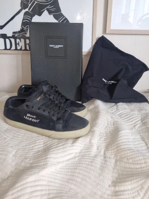 Saint luarent skor - !!ÄKTA!! Svarta sneakers från Saint Laurent med svart sula och guldfärgad logga på plösen. Skorna har låga skaft, snörning och är tillverkade i mocka med detaljer i läder. Snygg broderad logga på sidan. Påsen och lådan igår. kvitton finns och priset går att diskuteras! Skorna är perfekta för dig som vill ha en stilren och trendig look.