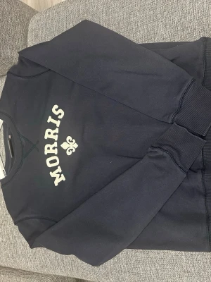 Mörkblå sweatshirt från Morris - Cool mörkblå sweatshirt från Morris med vit broderad logotyp och en liten fleur-de-lis på bröstet. Tröjan har rund hals, ribbade muddar och är tillverkad i mjukt material som är perfekt för en avslappnad look.