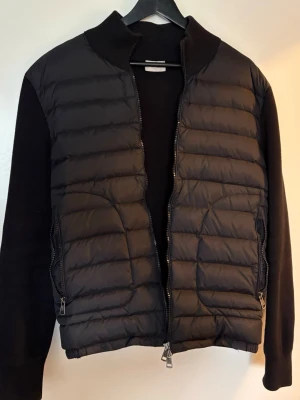 Svart cardiganjacka från Moncler - Pris går absolut att diskutera.Snygg svart cardiganjacka från Moncler med quiltad front och stickade ärmar. Jackan har dragkedja framtill, två sidofickor med dragkedja och Moncler-logga på ärmen. Perfekt för lager-på-lager och en clean look.