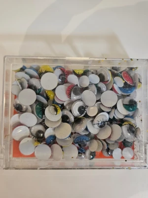 Färgglada rörliga plastögon - En ask med blandade rörliga plastögon i olika färger som grönt, gult, blått och rött. Perfekta till maskerad, cosplay eller pyssel. Ögonen har svarta fransar och kommer i olika storlekar. Lätta att limma fast på masker eller dräkter.