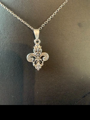 Silverfärgat halsband med lilja-hänge - Snyggt halsband i silverfärg med ett detaljerat liljaformat hänge. Kedjan är tunn och smidig, och hänget har en klassisk, ornamenterad design som ger en cool och tidlös vibe. Perfekt accessoar för att lyfta din stil.