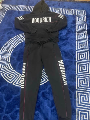 Svart HOODRICH loungewear set - Svart loungewear set från HOODRICH med matchande hoodie och byxor. Setet har vita logotryck på bröst, ärmar och ben samt tunna röda linjer längs benen. Hoodien har huva och byxorna har dragsko i midjan för skön passform.