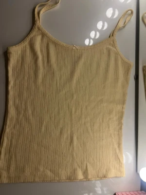 Gul ribbad linnetopp Brandy Melville - Söt ljusgul linnetopp från Brandy Melville i 100% bomull. Toppen har smala axelband och ett ribbat mönster med små håldetaljer. Perfekt för varma dagar och enkel att styla med jeans eller kjol.