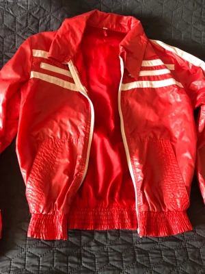 Röd retro vindjacka med vita ränder stl XS/ XXS - Cool klarröd vindjacka med vita ränder på bröstet och ryggen. Jackan har dragkedja framtill, elastiska muddar och krage. Materialet är syntetiskt, perfekt för dig som gillar retrostil och sportiga vibbar. Barn-stl eller en liten, späd vuxen person. Dragkedjan fungerar inte särskilt bra, behöver nog lagas.