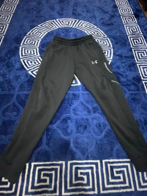 Svarta mjukisbyxor från Under Armour - Säljer ett par svarta mjukisbyxor från Under Armour med smal passform, resår i midjan och dragkedjeförsedd ficka på sidan. Byxorna har logga på låret och är perfekta för chill eller träning. Materialet känns mjukt och stretchigt.