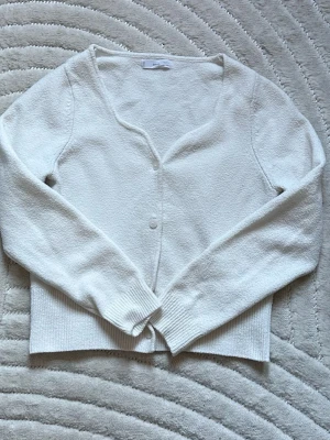 Vit kofta med v-ringning - Klassisk vit kofta med v-ringning och två knappar framtill. Långärmad modell med ribbade muddar och nederkant. Perfekt att slänga över en topp för en clean look. Materialet känns mjukt och lätt, vilket gör den skön att bära.