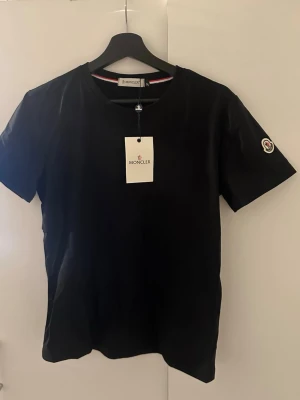 Svart Moncler t-shirt i bomull - Snygg svart t-shirt från Moncler med klassisk rund halsringning och korta ärmar. T-shirten har Moncler-logga på ärmen och är tillverkad i mjuk bomull. Perfekt för en stilren och enkel look.