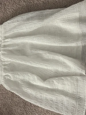 Vit kortkjol - Säljer en vit kortkjol med resår i midjan och ett diskret strukturerat mönster. Kjolen är gjord i luftig bomull och har en lätt volangig form som ger en avslappnad och trendig look.