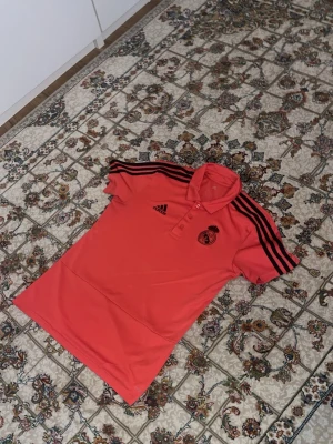 Röd Real Madrid piké från Adidas - Snygg röd pikétröja från Adidas med Real Madrid-logga på bröstet och svarta detaljer. Tröjan har klassisk krage, tre knappar och svarta ränder längs axlarna. Perfekt för dig som älskar fotboll och vill ha en sportig look.