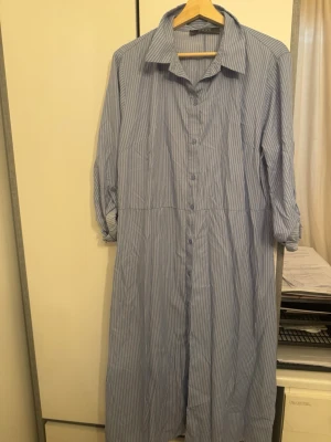 Blåvit randig skjortklänning Primark - Säljer en lång blåvit skjortklänning från Primark med tunna vertikala ränder. Klänningen har knappar hela vägen framtill, klassisk krage och långa ärmar som kan vikas upp. Perfekt för en avslappnad och stilren look.