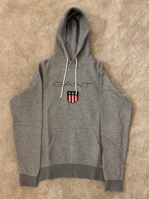 Grå hoodie från GANT  - Snygg grå hoodie från GANT med broderad logga och sköld på bröstet. Klassisk modell med huva och vita dragsnören samt känguruficka framtill. Mjuk bomullsblandning och ribbade muddar vid ärmslut och nederkant.