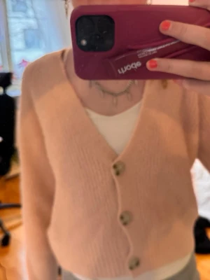 Ljusrosa kofta med stora knappar - Mysig ljusrosa kofta med v-ringning och tre stora knappar framtill. Koftan har en croppad passform och är stickad i ett mjukt material. Perfekt att slänga över en topp för en avslappnad look.