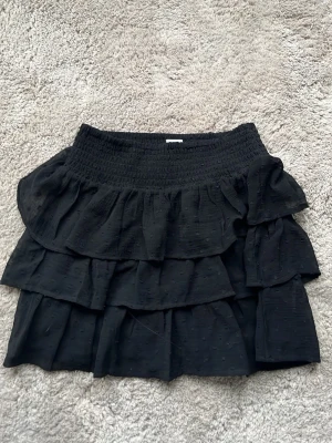 Svart volangkjol med resårmidja - Supersöt svart kortkjol med flera lager volanger och bred smockresår i midjan. Kjolen har en lätt struktur i tyget och ger en flowy känsla. Perfekt att styla med en enkel topp eller hoodie för en chill look. Finns en pytte liten fläck man inte ser och kanske något annat liknade. Men går bort i tvätt. Barn stolek!
