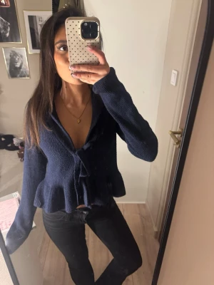 stickad tröja med volang och knyte - Snygg mörkblå stickad topp med volangkant och knytning framtill. Modellen har långa ärmar och en djup v-ringning. Perfekt för dig som vill ha en trendig och bekväm look. Materialet är mjukt och stretchigt.