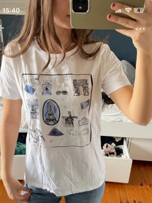 Vit t-shirt med Paris-motiv - Vit t-shirt med tryck av olika Paris-inspirerade motiv som Eiffeltornet, solglasögon och kameror. T-shirten har rund hals och korta ärmar. Perfekt för dig som gillar grafiska tryck och vill ha en enkel men snygg look.