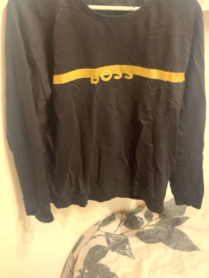 Svart BOSS sweatshirt med guld tryck  - Svart sweatshirt från BOSS med en guldig rand och BOSS-logga över bröstet. Tröjan har rund halsringning och långa ärmar. Passar till alla tillfällen