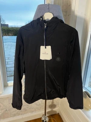 Moncler nylon vindjacka - Snygg svart vindjacka från Moncler med huva och dragkedja framtill. Jackan har en bröstficka med knapp och Moncler-logga, samt elastiska muddar vid ärmslut och nederkant. Tillverkad i lätt, glansigt material som står emot vind.