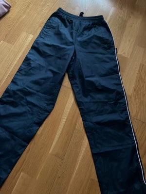 Svarta täckbyxor med vit rand - Säljer ett par svarta täckbyxor med vit rand längs sidorna och resår i midjan. Byxorna har rak passform och är tillverkade i ett slitstarkt, vattenavvisande material. Perfekta för kalla dagar när du vill hålla dig varm och torr.