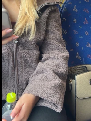 Brun teddyjacka med dragkedja - Mysig brun teddyjacka med dragkedja framtill och hög krage. Jackan har en fluffig och mjuk textur som ger en avslappnad look. Perfekt för kyliga dagar och enkel att matcha med olika outfits.