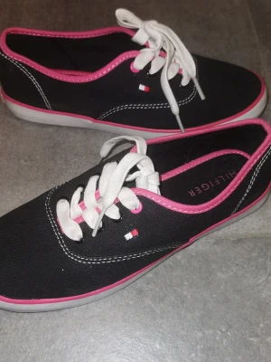 Svarta Tommy Hilfiger sneakers,  - Snygga svarta sneakers från Tommy Hilfiger med vita snören och rosa detaljer runt kanten. Skorna har en låg profil, vit sula och den klassiska Hilfiger-loggan på sidan. Tillverkade i canvasmaterial för en avslappnad och trendig look. Super snygga, de hae använts vid ett fåtal gånger, har några mindre märken som går säkert bort med lite vatten (se bilder) , i övrigt mycket fint skick. Köpta utomlands, storlek 36