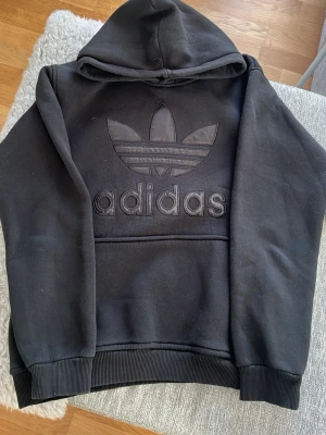 Svart hoodie från Adidas Originals - Svart hoodie från Adidas Originals med stor broderad Trefoil-logga och adidas-text på bröstet. Klassisk känguruficka framtill, ribbade muddar och huva. Tillverkad i mjukt sweatshirtmaterial som känns skönt mot huden. Har även ett par matchande mjukis byxor till ( kostar tillsammans 400kr )