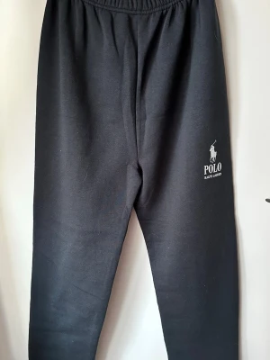 Svarta mjukisbyxor Polo Ralph Lauren - Svarta mjukisbyxor från Polo Ralph Lauren med vit logga tryckt på vänster ben. Byxorna har resår i midjan och är gjorda i mjukt, bekvämt material. Perfekta för chill dagar eller när du vill ha en avslappnad stil.