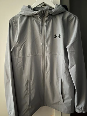 Ljusgrå vindjacka från Under Armour - Snygg ljusgrå vindjacka från Under Armour med huva, dragkedja framtill och två fickor. Jackan har logga på bröstet och stort tryck på ryggen. Perfekt för blåsiga dagar och sportiga outfits.