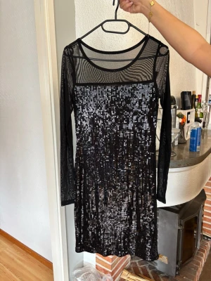 Svart paljettklänning med meshärmar - Snygg svart kortklänning med långa meshärmar och glittriga paljetter framtill och baktill. Klänningen har rund halsringning och är perfekt för dig som vill sticka ut. Materialet är polyester och mesh, vilket ger en cool och trendig look.