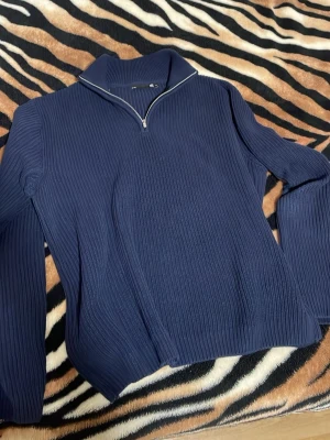 Mörkblå stickad tröja med halvzip - Mörkblå stickad tröja med ribbad struktur och halv dragkedja vid kragen. Tröjan har lång ärm och hög krage, perfekt för lager-på-lager. Enkel och stilren design som passar till många outfits.